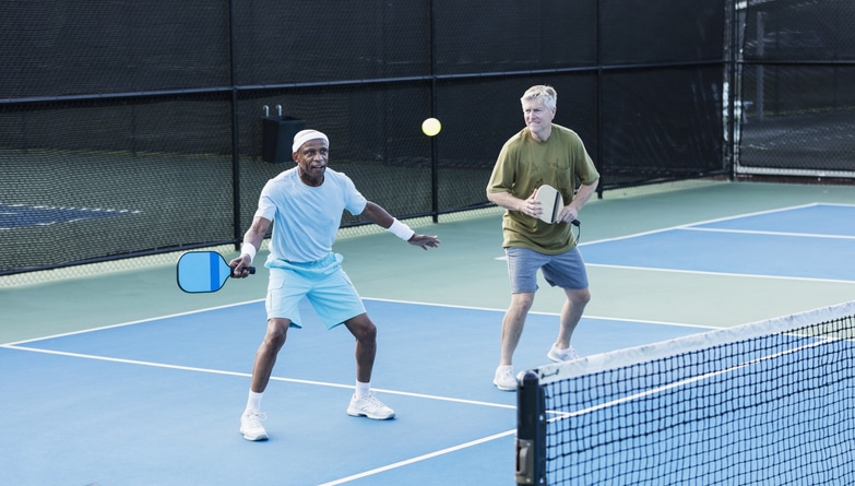 Two multi-ethnic senior  men playing paddleball. They are a doubles team. The African-American man, in his 70s, is concentrating as he reaches to hit the ball with his paddle.  His partner, in his 60s, is watching and smiling.
