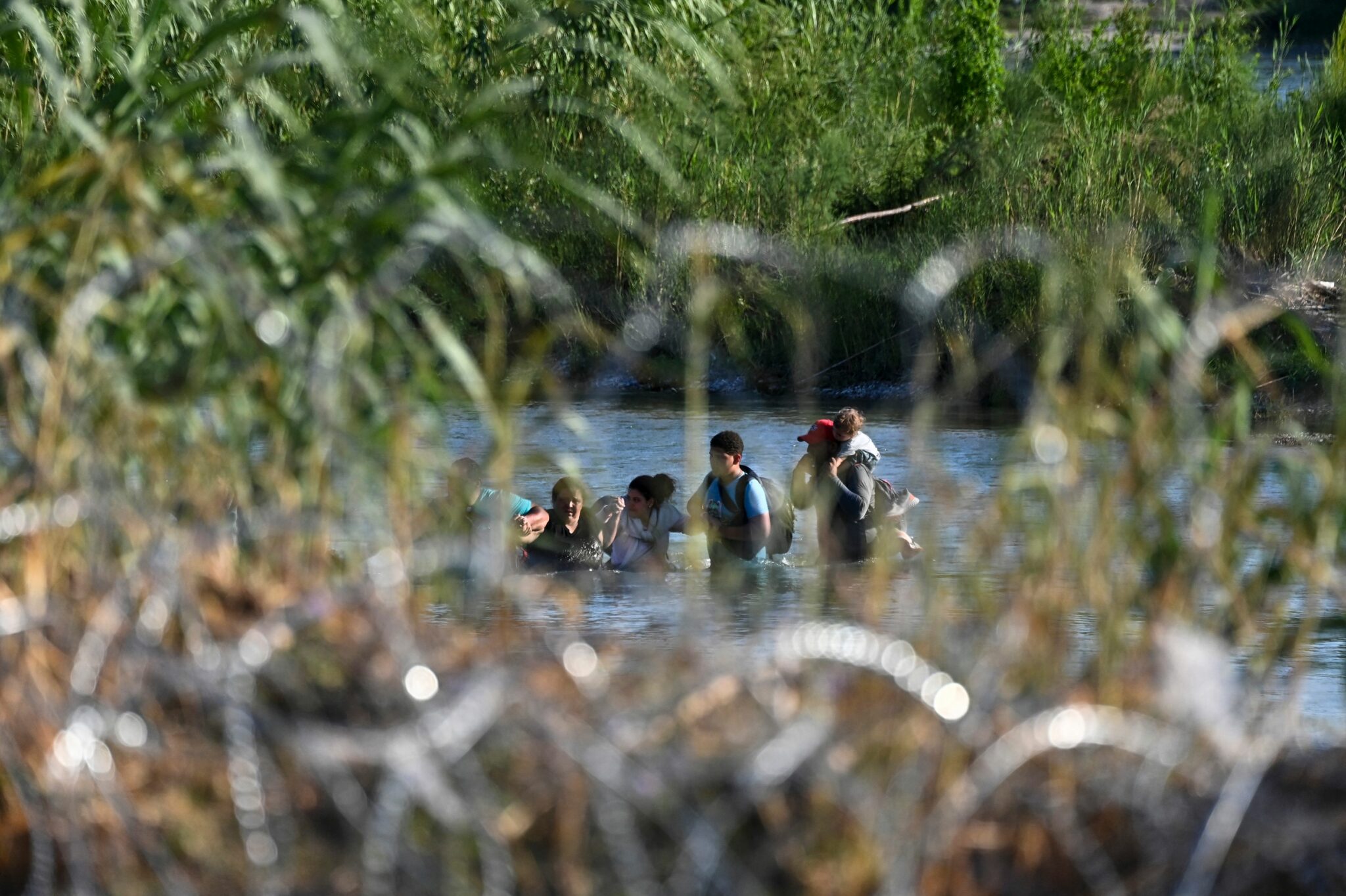 TOPSHOT - A migrant family from Venezuela is seen beyond concertina wire as they illegally cross the Rio Grande River in Eagle Pass, Texas, at the border with Mexico on June 30, 2022. - Every year, tens of thousands of migrants fleeing violence or poverty in Central and South America attempt to cross the border into the United States in pursuit of the American dream. Many never make it. On June 27, around 53 migrants were found dead in and around a truck abandoned in sweltering heat near the Texas city of San Antonio, in one of the worst disasters on the illegal migrant trail. (Photo by CHANDAN KHANNA / AFP) (Photo by CHANDAN KHANNA/AFP via Getty Images)